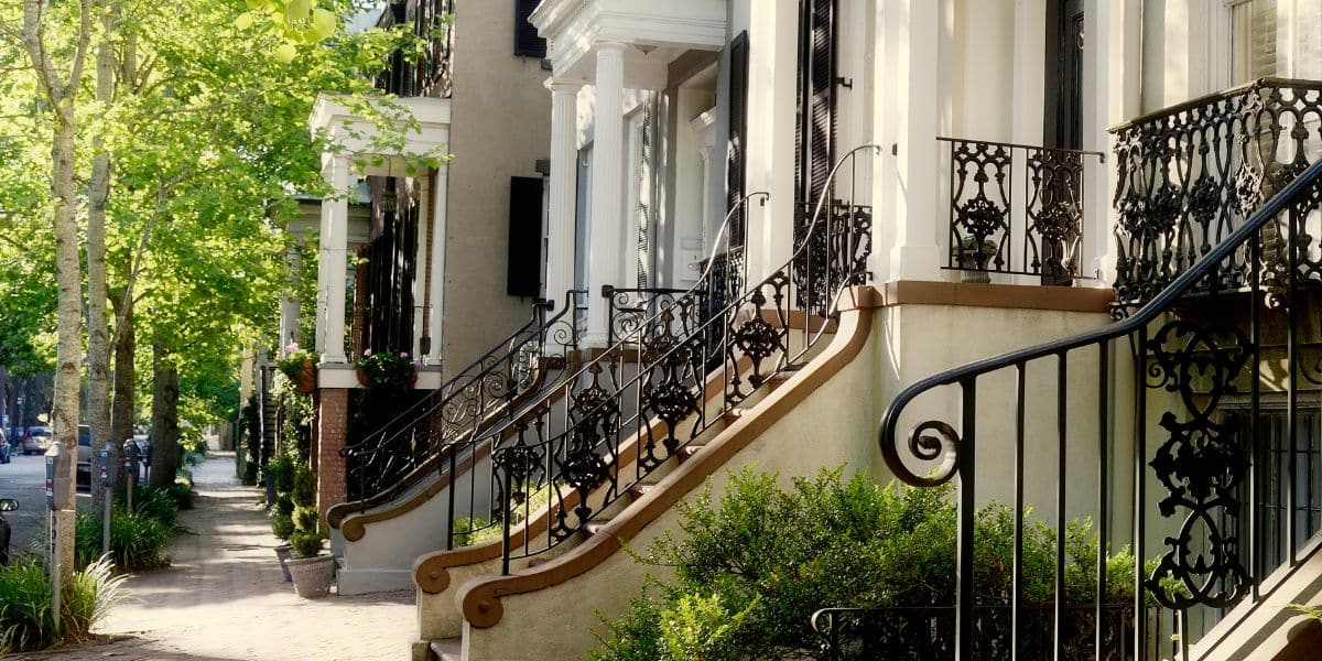 Historic downtown Savannah row homes with ornate iron railings, columned porches, and tree-lined sidewalks in bright morning light.